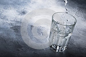 Last drops of pure water are poured into large glass cup on gray table background, selective focus