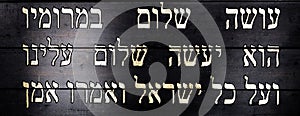 Last lines of Jewish prayer Kaddish on black wooden background. Magnification and sanctification of God`s name