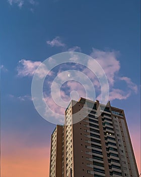 Late afternoon with blue and pink sky with clouds between the moon next to tall buildings in the city