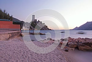 Late evening view of Castle of Malcesine with moving sea water, trees & boat at Paina Beach, Lake Garda, northern Italy
