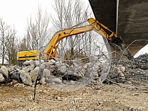 Lateral view of an excavator running a reinforced concrete demolition with a hydraulic breaker under a bridge under construction