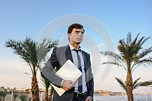 Lateral view of a young man in suit with laptop working on green grass near a resort