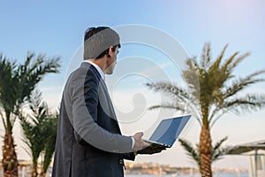 lateral view of a young man in suit with laptop working on green grass near a resort