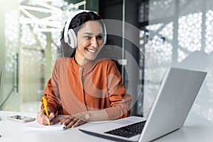 Latin american business woman with curly hair and headphones watching online training course at workplace, woman writing