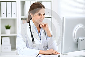 Latin american female doctor sitting at the table and working by computer at hospital office. The physician or therapist