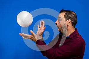 Latin bearded man dressed in a purple shirt with a cork ball isolated on blue studio background, he is throwing the ball