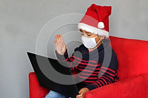 Latin boy with protection mask and santa claus hat in video call with laptop, new normal covid-19 and christmas decoration