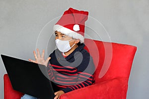 Latin boy with protection mask and santa claus hat in video call with laptop, new normal covid-19 and christmas decoration