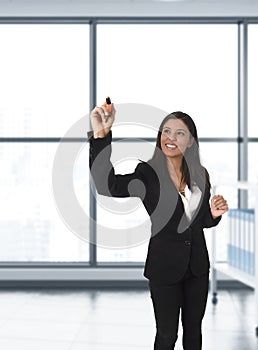 Latin business woman in formal suit writing with marker on invisible virtual screen or board at modern office