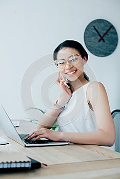 Latin businesswoman talking on smartphone and using laptop at workplace