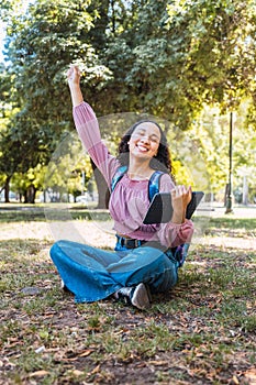 Latin college student woman celebrating and holding a tablet sitting outside in a park on the grass