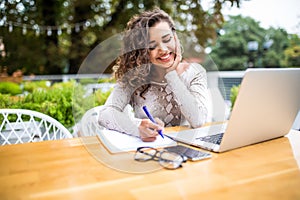 Latin curly Woman typing working on laptop on wooden table and making notice at notebook working on laptop while lunch break in ca