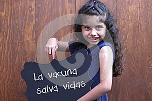 Latin little girl with curly hair shows her arm recently vaccinated against Covid-19 with a black sign that says `The vaccine save