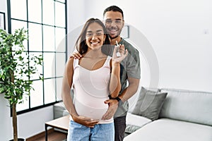 Latin man and woman couple hugging each other holding key of new house expecting baby at home
