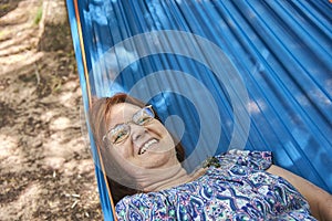 Latin mature woman on vacations wearing glasses smiling lying in a blue hammock