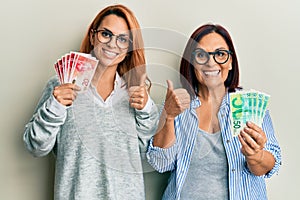 Latin mother and daughter holding israel shekels smiling happy and positive, thumb up doing excellent and approval sign