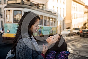 Latin mother and daughter looking affectionately outside on a Lisbon street, in the background there is a classic tram from