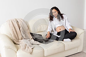 Latin pregnant woman using laptop sitting on sofa at home. Pregnancy and information for parenthood concept.