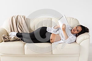 Latin pregnant woman using tablet computer while lying on sofa at home. Pregnancy and information for parenthood concept