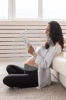 Latin pregnant woman using tablet computer sitting near sofa at home. Pregnancy and information for parenthood concept.