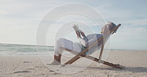 Latin senior hispanic woman relaxing on sunbed on beach at sunset, reading book