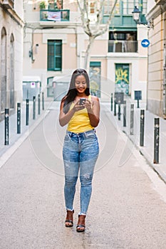 Latin tourist girl with travel suitcase is walking on the street and using a mobile phone to book a hotel