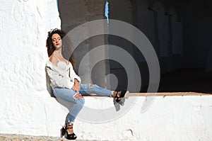 Latin woman with dark curly hair and middle age is sitting on a wall. The woman is sad and pensive. The woman is a single mother