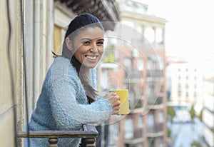 Latin woman drinking cup of coffee or tea smiling happy at apartment window balcony