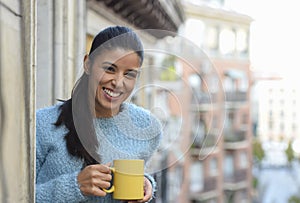 Latin woman drinking cup of coffee or tea smiling happy at apartment window balcony