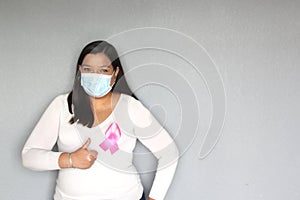 Latin woman with protection mask, black hair and white sweater with pink ribbon for campaign against breast cancer