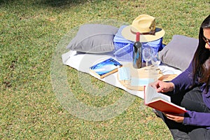 Latin woman reading book sitting, with picnic basket, wine and food outdoors in the forest