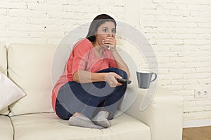 Latin woman sitting at home sofa couch in living room watching television scary horror movie