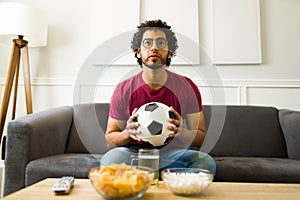 Latin young man looking worried while watching a soccer game on tv