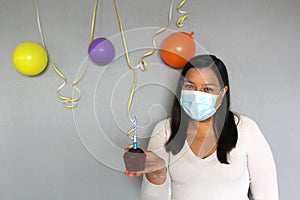 Latina with black hair with protection mask, cake, colored balloons celebrates birthday at home by quarantine and new normal covi