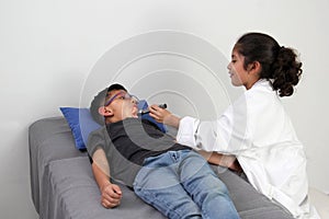 Latino girl and boy play to be a doctor with a white coat that is too big for her and patient who acts to feel sick very entertain