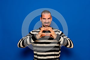 Latino man with beard putting his hands together diabolically showing his evil intentions, isolated on blue studio