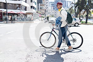 Latino man in casual clothes walking down the street with his bike wearing a red cap, yellow headphones and a backpack in the city
