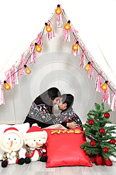 Latino mom and son celebrate Christmas under a teepee dressed in ugly sweaters with lights and Christmas decorations show their lo