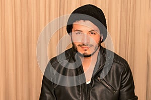 Latino young adult man in leather jacket wearing polyester hat with a happy expression with a beige curtain background