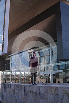 Latino young man dressed in urban clothing posing on a high fence with architecture in the background