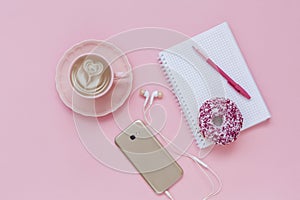 Latte Coffee cup, delicious pink donut with sprinkle, notebook and pen, smart phone with headphones on pink paper background