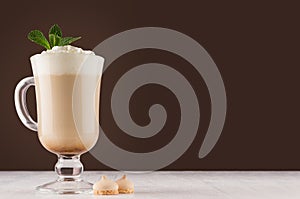 Latte coffee in elegant transparent glass with foam, fresh mint and biscuits in modern dark brown kitchen interior, copy space.
