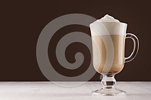 Latte coffee in elegant transparent glass with foam in modern dark brown kitchen interior, copy space.