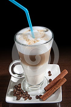 Latte macchiato in a glass with white plate, spoon, decorated with coffee beans, cinnamon. Black background.