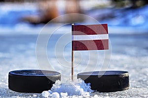 Lotyšská vlajka na párátku mezi dvěma hokejovými puky. Lotyšsko bude hrát na mistrovství světa ve skupině B. 2019 IIHF World Championship