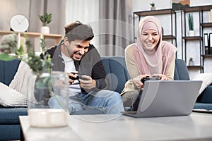 Laughing 30-aged funny Muslim couple having fun together while playing video games at home using gamepads. Family