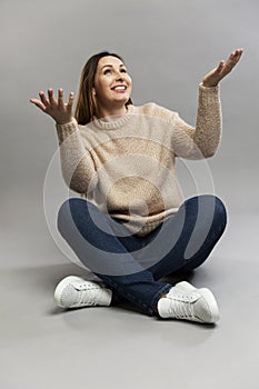 Laughing adult woman sits on the floor with her legs crossed and her arms raised up. Beautiful brunette in jeans and a beige