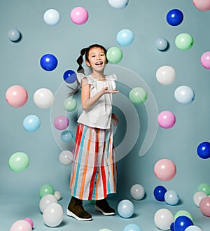 Laughing asian kid girl in party clothes is having fun surrounded by colorful air balloons holds hand up with open palm