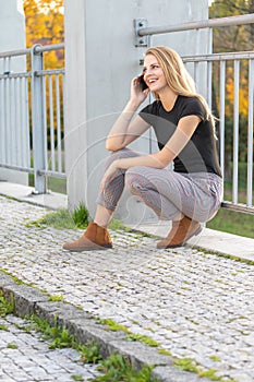 Laughing attractive young girl is squatting and phoning on the bridge. Vertically