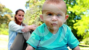 Laughing baby boy sitting on the grass with mother behind him